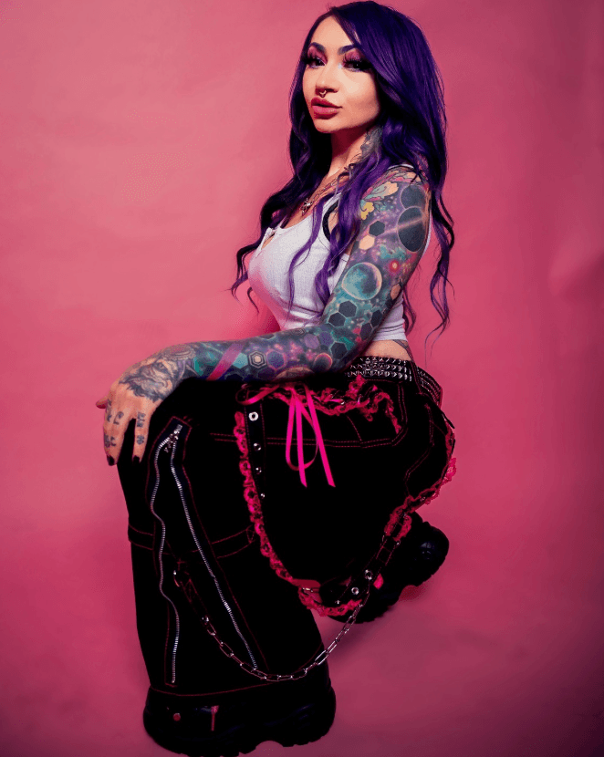 Courtney Raimondi kneeling in front of a pink backdrop, showing colorful tattoo work.